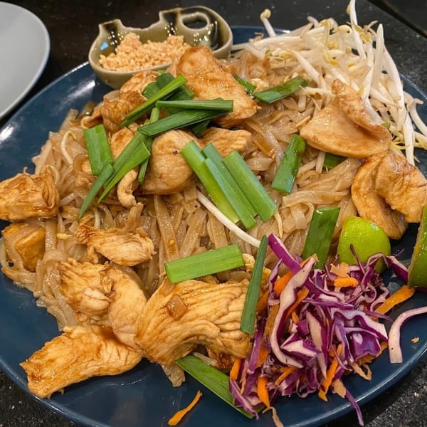 Chicken Pad Thai with bean sprouts, green onions, shredded cabbage, carrots, and lime on a blue plate.